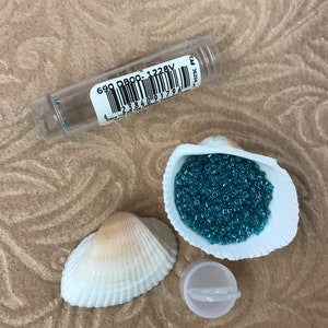 May include: A small plastic container of teal blue seed beads, a seashell filled with the beads, and a second seashell. The container has a barcode and the text "690 DB00-1228V".