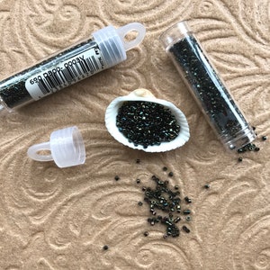 May include: Two small clear tubes filled with black iridescent seed beads. The beads are also shown in a white seashell and scattered on a brown surface. The tubes have a barcode label with the text "690 D80P 0003V".