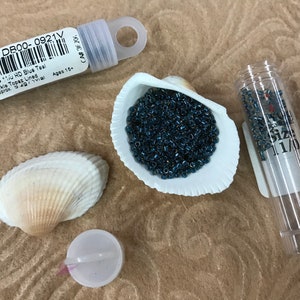 May include: A small clear tube filled with dark blue seed beads. The tube has a label that reads "S90 DB00-0921V Delica 11/0 HD Blue Teal Sparkle Topaz Lined Approx 3.4g/vial Ages 15+". The beads are also displayed in a white seashell.