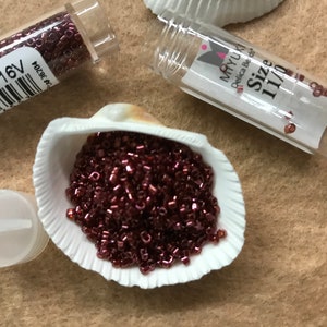 May include: A small white seashell filled with a pile of small, dark red seed beads. The beads are in a clear plastic tube labelled "MIYUKI Delica Beads Size 11/0".