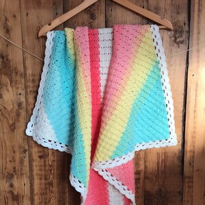 May include: A rainbow crochet baby blanket with a white crocheted border. The blanket is hanging on a wooden hanger.