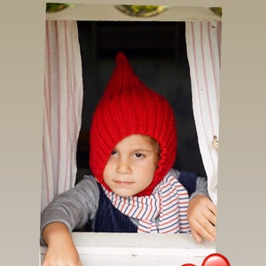 May include: A child wearing a red knitted hat with a pointed top looks out of a window. The window is framed by white wood and has pink and white striped curtains. The text "Cozy & warm" is above the window. A red heart is in the bottom right corner of the image.