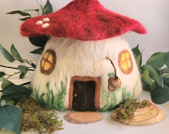 May include: A felt mushroom house with a red roof, white walls, and a brown door. The house has two yellow windows and a small acorn hanging from the roof. The house is surrounded by green felt grass and moss.