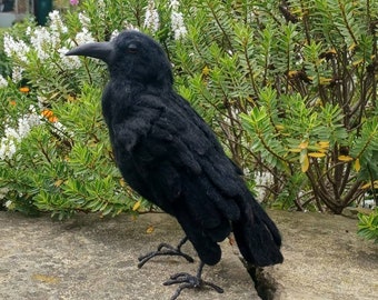 May include: A black crow figurine perched on a gray stone surface. The crow is made of felt and has a realistic appearance. The background features a green bush with white flowers.