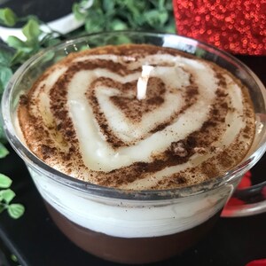 May include: A glass candle shaped like a coffee mug with a white candle and brown cinnamon sprinkles on top. The candle has a heart shape in the center.