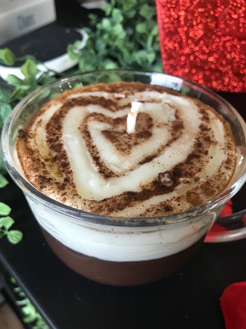 May include: A glass candle shaped like a coffee mug with a white candle and brown cinnamon sprinkles on top. The candle has a heart shape in the center.