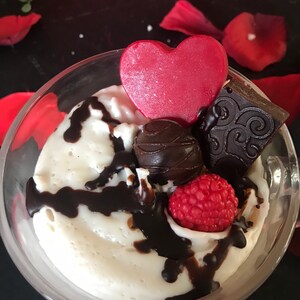 May include: A clear glass dessert dish filled with a white mousse topped with chocolate sauce, a chocolate heart, a chocolate truffle, a red raspberry, and a chocolate square with a decorative design.