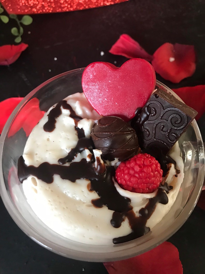 May include: A clear glass dessert dish filled with a white mousse topped with chocolate sauce, a chocolate heart, a chocolate truffle, a red raspberry, and a chocolate square with a decorative design.