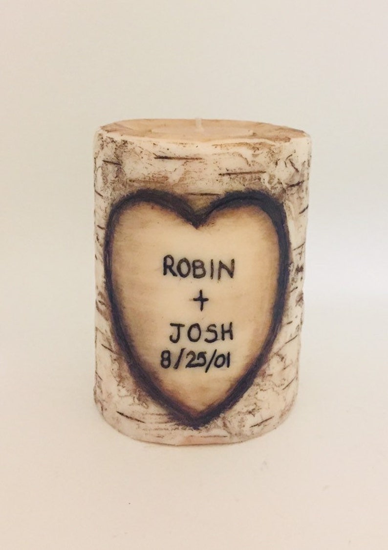May include: A white cylindrical candle with a birch bark design. A heart is carved into the candle with the names "Robin + Josh" and the date "8/25/01" inscribed inside.