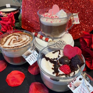 May include: Three glass candle jars with a latte base and different toppings. The first jar has a red hot cinnamon bun latte, the second jar has a raspberry chocolate drizzle latte, and the third jar has a vanilla latte with pink sprinkles. The jars are on a red glitter surface with red rose petals.