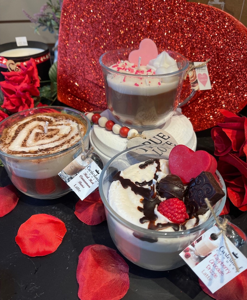 May include: Three glass candle jars with a latte base and different toppings. The first jar has a red hot cinnamon bun latte, the second jar has a raspberry chocolate drizzle latte, and the third jar has a vanilla latte with pink sprinkles. The jars are on a red glitter surface with red rose petals.