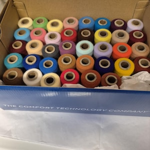 May include: A cardboard box filled with colorful spools of thread. The spools are arranged in neat rows and feature a variety of colors, including blue, green, yellow, pink, and orange. The box has the text "THE COMFORT TECHNOLOGY COMPANY" printed on the side.