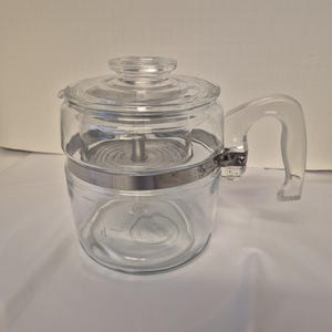 May include: A clear glass coffee percolator with a glass lid and handle. The percolator has a stainless steel band and a filter basket inside. The handle is clear and curved. The percolator is designed to brew up to 4 cups of coffee.