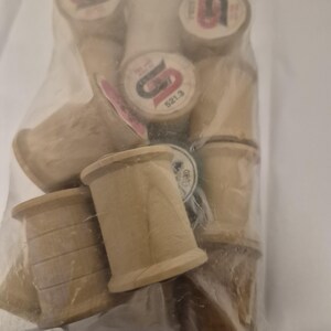 May include: A clear plastic bag filled with several wooden spools. The spools are light brown with a cylindrical shape and a slightly flared base. Some spools have labels with red and black text. The spools are of various sizes.