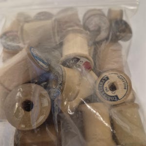 May include: A clear plastic bag filled with a collection of vintage wooden spools. The spools vary in size and color, with some showing remnants of thread and labels. The spools are a light brown color.