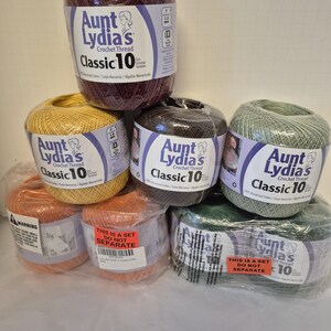 May include: A collection of Aunt Lydia's Classic 10 crochet thread balls in various colors including burgundy, yellow, dark brown, sage green, and orange. Each ball features a white label with blue text and a graphic of a woman's profile.