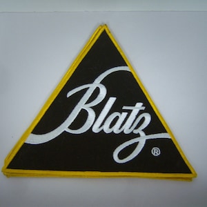May include: Black triangular patch with the word "Blatz" in white script. The patch has a yellow border and is likely an embroidered logo.