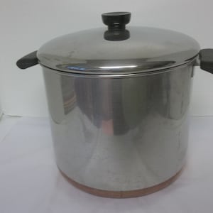 May include: A stainless steel cooking pot with a lid and two side handles. The pot has a copper-colored base and a black handle on the lid. The pot is designed for cooking and food preparation.