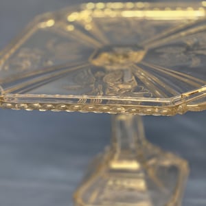 May include: A clear, octagonal glass cake stand with a detailed, embossed design. The stand features a pedestal base and a decorative beaded edge. The glass has a slight amber tint, and the design includes radial patterns and floral motifs.