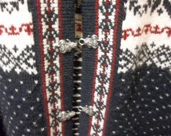 May include: Close-up of a gray, white, and red patterned sweater. The sweater features a detailed knit design with silver clasps. The overall style suggests a traditional, possibly Scandinavian, design.