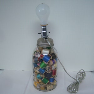 May include: A unique lamp made from a clear glass jar filled with colorful spools of thread. The jar is topped with a silver lid and a light fixture with a frosted bulb. A silver power cord extends from the lamp.