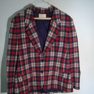 May include: A plaid blazer in red, white, and navy blue. The jacket has a two-button closure, notched lapels, and two front pockets. The interior lining is a deep purple color. The blazer is hanging on a wooden hanger.