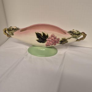 May include: A vintage ceramic centerpiece bowl with a pink interior and a white exterior, adorned with a grape cluster and leaf design. The bowl has gold-colored handles and sits on a green pedestal base. A decorative piece for the home.