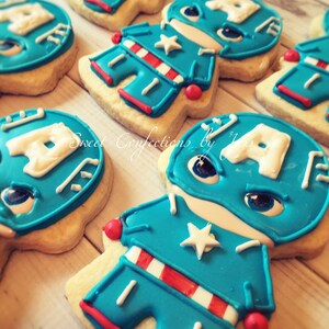May include: Decorated sugar cookies shaped like a superhero with a blue and red costume, a white star on the chest, and a white letter A on the head. The cookies are on a light wood surface.