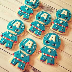 May include: Six decorated sugar cookies shaped like a superhero character. The cookies are blue, red, and white with a white letter "A" on the front. The cookies are on a light wood surface.