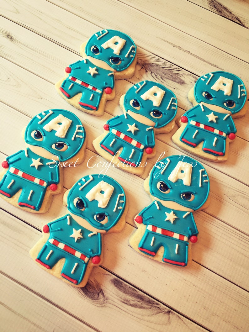 May include: Six decorated sugar cookies shaped like a superhero character. The cookies are blue, red, and white with a white letter "A" on the front. The cookies are on a light wood surface.