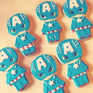 May include: Six decorated sugar cookies shaped like Captain America. Each cookie is blue, white, and red with a white star on the chest and a white letter 'A' on the head.