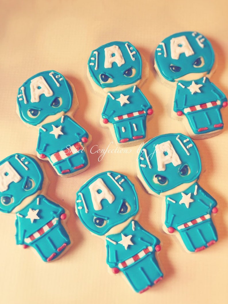 May include: Six decorated sugar cookies shaped like Captain America. Each cookie is blue, white, and red with a white star on the chest and a white letter 'A' on the head.