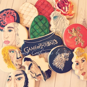 May include: A collection of Game of Thrones themed sugar cookies decorated with various characters and symbols from the show. The cookies are arranged on a wooden surface and include images of Daenerys Targaryen, Jon Snow, and the direwolf sigil.