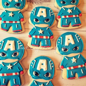 May include: Six decorated sugar cookies shaped like Captain America. Each cookie has a blue and red costume with a white star on the chest. The cookies are on a wooden surface.