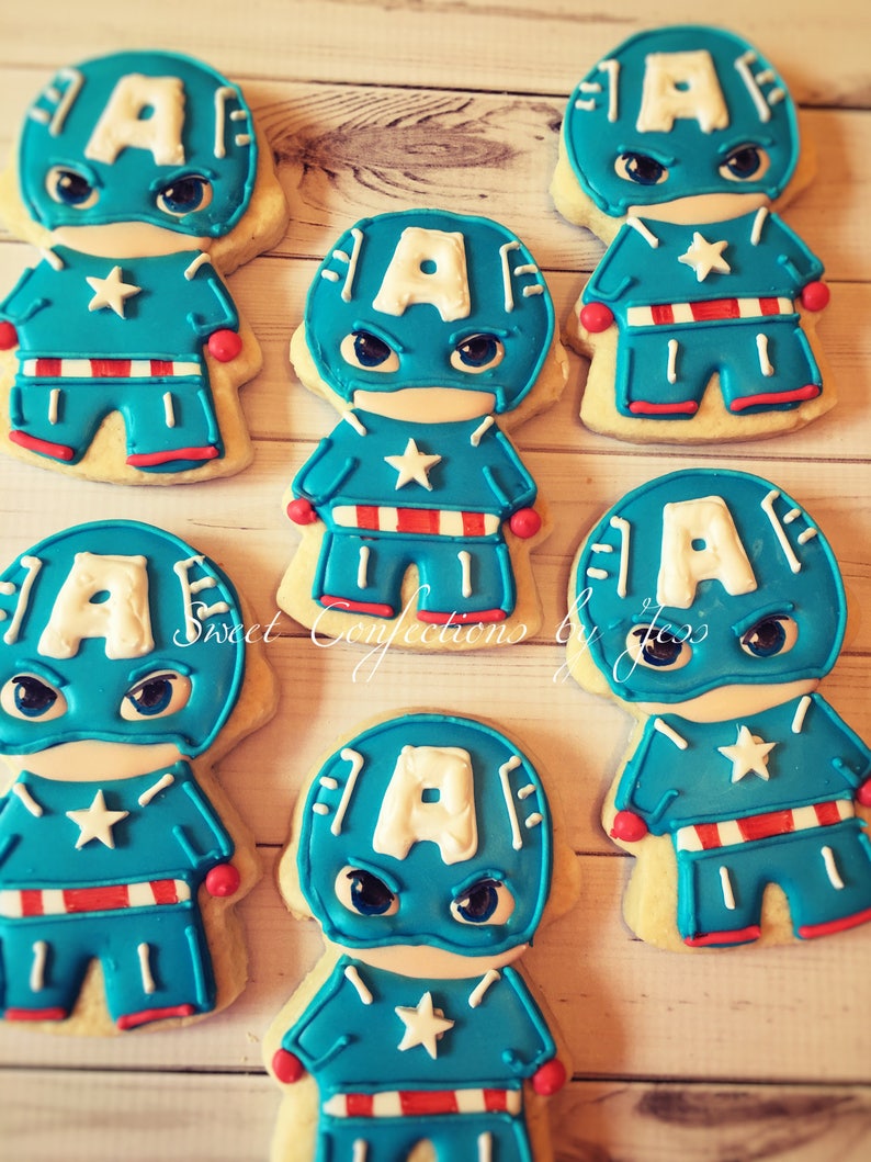May include: Six decorated sugar cookies shaped like Captain America. Each cookie has a blue and red costume with a white star on the chest. The cookies are on a wooden surface.