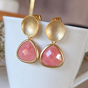 Könnte beinhalten: Ein Paar goldfarbene Ohrringe mit einem kreisförmigen, texturierten Oberteil, das mit einem facettierten, dreieckigen rosa Steinanhänger verbunden ist. Das Metall hat ein mattes Finish und die Steine sind in einem glatten Goldrahmen gefasst.