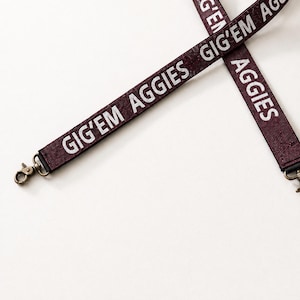 May include: A maroon lanyard featuring the white text GIG'EM AGGIES repeated along the fabric strap. The lanyard includes a metallic bronze-toned lobster claw clasp at the end for attaching items, with a textured finish on the maroon material.