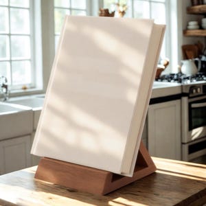 May include: A large, off-white book rests on a wooden book stand. The book has a light-colored cover and a thick spine. The stand is made of a warm-toned wood and has a triangular shape, designed to hold the book upright.