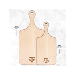 Puede incluir: Dos tablas de cortar de madera con un mango. La tabla grande mide 17,8 cm por 38,1 cm y la tabla pequeña mide 14 cm por 29,2 cm. Ambas tablas tienen las letras "A M" y una "T" estilizada en el centro.