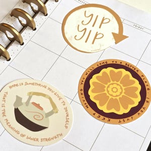 May include: Three circular stickers placed on a white planner page with gold binding. One sticker features the text 'yip yip' with an arrow. Another shows a teapot with the text 'Hope is something you give to yourself'. The third displays a flower.