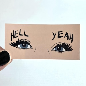 May include: A rectangular sticker with a light peach background featuring two stylized eyes with long black eyelashes and blue irises. The word HELL is printed in black above the left eye, and the word YEAH is printed in black above the right eye.
