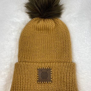 May include: Mustard yellow knit beanie with a dark brown faux fur pom-pom. The beanie features a brown leather patch with a yarn and needle design. This warm hat is perfect for cold weather and adds a stylish touch to any outfit.
