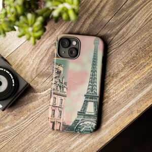 May include: A phone case with a vintage-style image of the Eiffel Tower and a building in Paris, France. The case has a pink and blue background with a faded look.