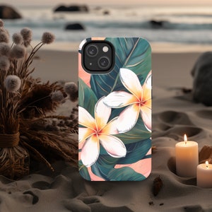 May include: A phone case featuring a floral design with white and yellow flowers, green leaves, and a pink background. The case is set on a sandy beach with candles and dried plants in the background. The case is designed for a smartphone.
