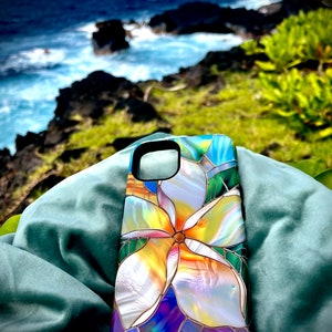 Puede incluir: Una funda de teléfono con un diseño de vitral colorido que presenta una flor de plumeria blanca. El fondo es una imagen borrosa de una costa rocosa y el océano.