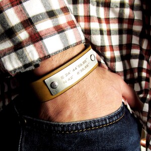 May include: A brown leather bracelet with a silver plate engraved with the coordinates "E 24° 44' 59.85" N 42° 8' 35.69"