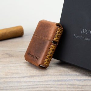 Op de afbeelding: Een bruine leren aanstekerhoes met een gestikt ontwerp en de tekst "Broscolors" gegraveerd op de voorkant. De hoes ligt op een houten oppervlak naast een sigaar en een zwarte doos met de tekst "Broscolors Handmade Leather Accessories" op de voorkant.