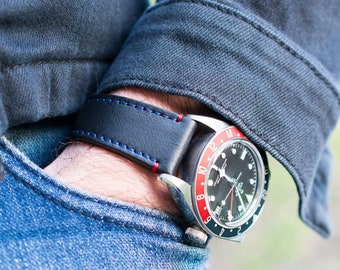 May include: A black leather watch strap with red and blue stitching. The watch has a black face with white markings and a silver bezel.