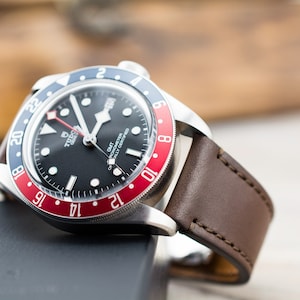 Puede incluir: Un reloj de pulsera con esfera negra y correa de cuero marrón. La esfera del reloj tiene un bisel rojo y azul con números blancos y una ventana de fecha. La esfera del reloj también tiene el texto "TUDOR" y "GMT" impreso en él.