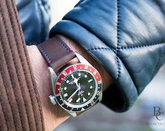 May include: A brown leather watch strap with blue stitching on a wrist wearing a silver watch with a black face and red and white numbers around the edge. The watch face has a date window at the 3 o'clock position and the text "ROLEX" is visible.